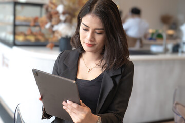 A beautiful career woman, wearing a black blazer, is holding a tablet in a cafe.
