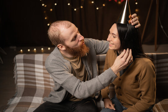 Young couple preparing to celebrate birthday party at home. Husband helping wife to put party hat on her head. Birthday preparation