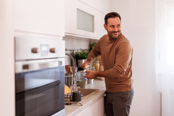 Confident mid adult man smiling and looking away while washing drinking glass in kitchen sink at home