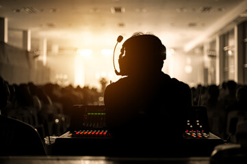Silhouette of a sound engineer wearing a headset, managing audio equipment during a live performance. Warm golden light illuminates the stage and the crowd, creating a dramatic event scene.