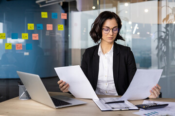 Confident businesswoman analyzing papers in office setting, demonstrating focus and determination. Modern workplace features laptop and charts, emphasizing productivity.