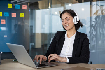 Woman in business suit smiling, typing on laptop at office desk with headphones. Engaged in online...