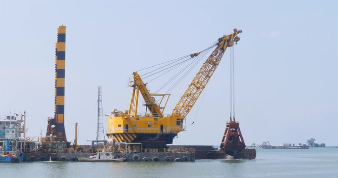 large, yellow floating crane on a barge in a body of water. The crane has a long boom with multiple cables and is equipped with a clamshell bucket, used for dredging.