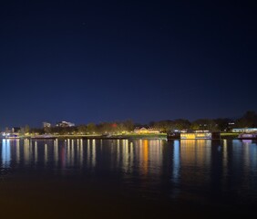 A serene riverside view at night, featuring illuminated buildings and trees reflected on the calm water, with a clear starry sky in the background.