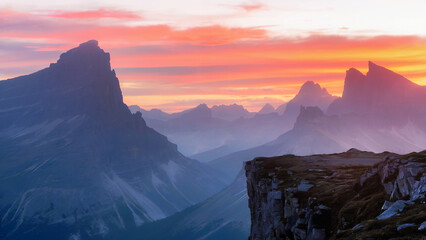 A breathtaking landscape during sunset or sunrise is enhanced by the presence of towering mountain peaks, with one particularly sharp and jagged peak standing out prominently.