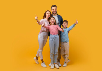Fototapeta premium Happy family consisting of a mother, father, son, and daughter stands close together, all giving a thumbs up sign. They are posing against a vivid yellow backdrop