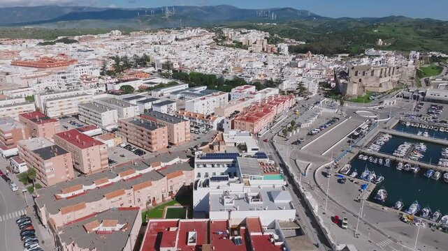 Aerial view of Tarifa's white buildings, Guzman Castle, and wind turbines. The Atlantic Ocean and distant mountains frame the Andalusian town.
