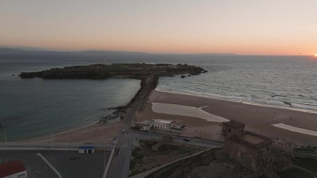 Aerial view of Tarifa with a lighthouse on the peninsula, historic buildings, and the Atlantic Ocean at sunset. Morocco is visible across the strait.