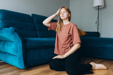 Obraz premium Blonde 30s woman has a headache, migraine. Young girl in bad thoughts feeling lonely indoors and touching her head with her eyes closed and sitting on floor near a sofa at home