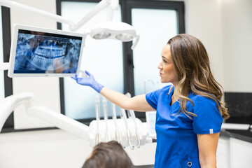 A female dentist in blue scrubs stands in a clinic, pointing at X-rays on a screen, as she explains...