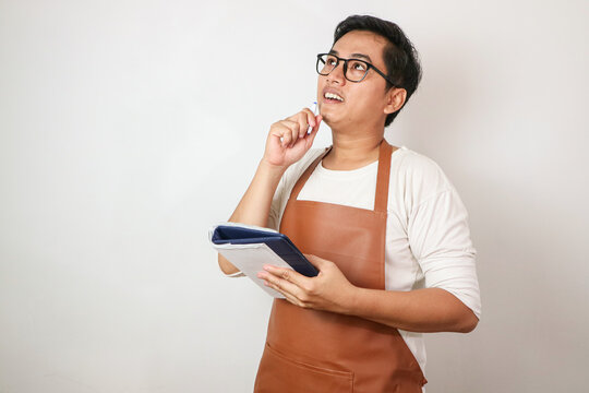 Asian barista man in brown apron taking order, writing on menu book list, thinking with pen on head. Isolated image on white background