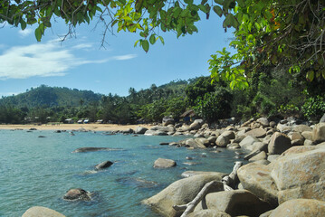 A serene tropical beach scene featuring a rocky shoreline, clear blue water, and lush greenery under a bright sky.