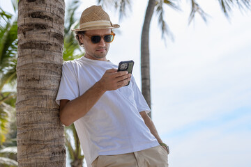 Handsome attractive man with sunglasses brimmed hat white T-shirt using smartphone chatting messaging on tropical beach standing under palm tree in vacation summer travel.