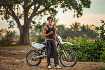 Fototapeta premium Young Hispanic man in black tank top smiling confidently next to white dirt bike on rural trail during golden hour, tropical trees in background