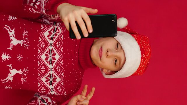 happy Caucasian boy 9-10 years old in Santa hat and New Year red sweater makes selfie on phone camera, vertical video, red studio background