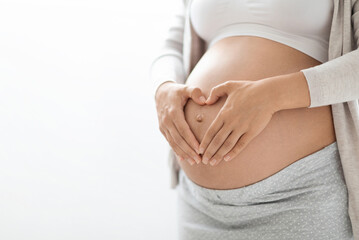 Cropped shot of pregnant woman in homewear holding hands in heart-shaped symbol gesture on big...