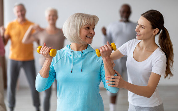 Female fitness coach helping active senior woman while exercising with dumbbells, sporty multiracial group of elderly people having fitness class at nursing home, training with instructor