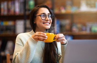 Benefits of remote work. Dreamy lady enjoying coffee during work on laptop computer in city cafe,...