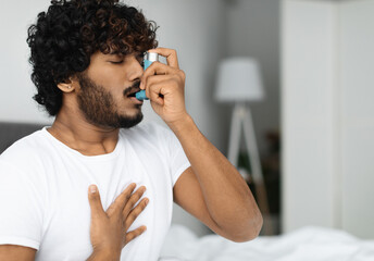 Indian guy suffering from asthma, using inhaler in bed, side view, copy space. Young hindu man woke...