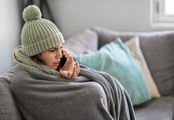 Stressed Young Woman Talking On Cellphone While Sitting On Couch Wrapped In Blanket, Upset Female...