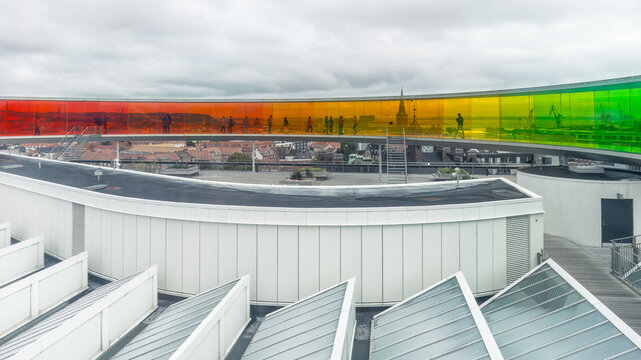 Panoramic view of the rooftop rainbow walkway at the ARoS Aarhus Art Museum with colorful glass panels and cloudy sky in the background. Motion blurred people. Denmark