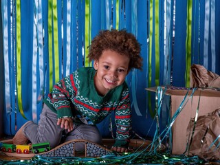 Smiling Multi ethnic boy at Christmas, wearing a festive jumper sits with his presents. Blue and green glitter backdrop.