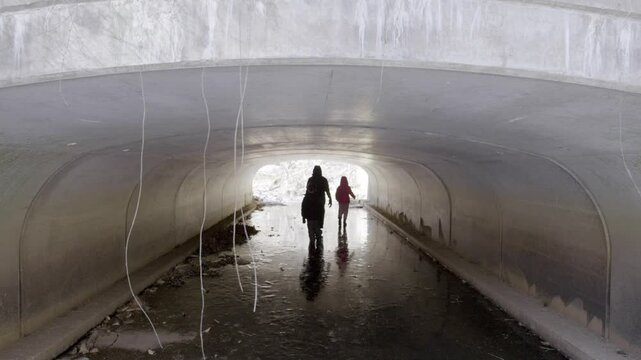 Three children in snow outfits walking along a frozen creek under a culvert. One of them kicks a rock along the ice. In Chapel Hill, North Carolina in winter.