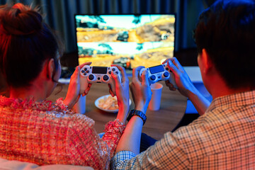 Couple gamer with controlled joystick playing car racing video game together on tv screen with snack at back side view, sitting sofa at neon light colorful living room at modern comfy home. Infobahn. © InfiniteFlow