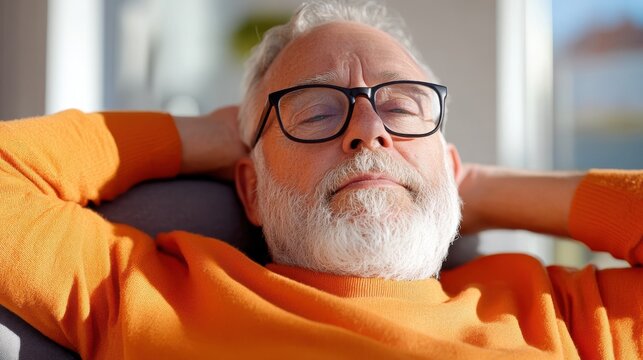 A senior man with glasses and a beard leans back with his hands behind his head, appearing relaxed and content, wearing a bright orange sweater indoors.