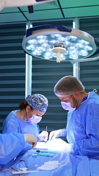 Medics perform surgery on thyroid. Chief female surgeon and assisting doctor work under bright round lamp. Vertical video.