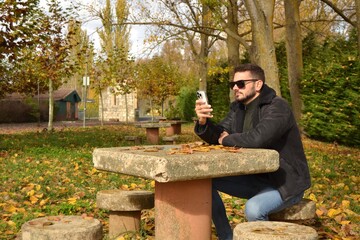 Fototapeta premium Young handsome guy walking through the park or forest enjoying the autumn. New technologies, mobile phone. Looking for coverage. Taking advantage of free time to read. E-book.Reconnecting with oneself