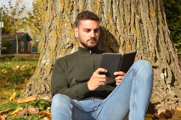 Fototapeta premium Young handsome guy walking through the park or forest enjoying the autumn. New technologies, mobile phone. Looking for coverage. Taking advantage of free time to read. E-book.Reconnecting with oneself