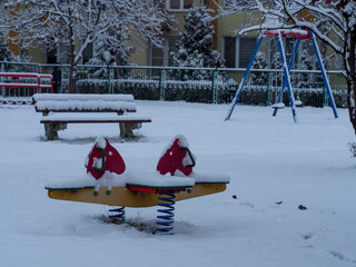 Snowy playground with a seesaw, bench, and swing set covered in snow, creating a serene winter scene