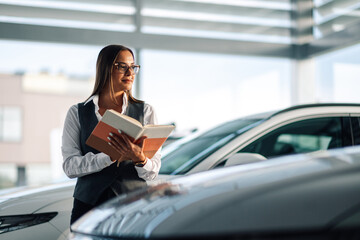 Stylish woman holding notebook near vehicle in a car dealership