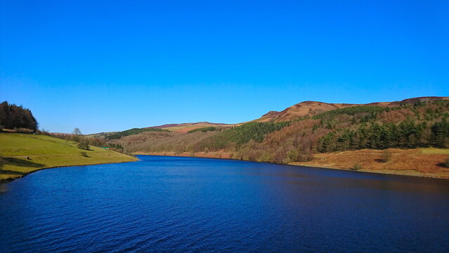 Peak District, UK - 2.25.2018: A peaceful and tranquil scene of calm blue Ladybower Reservoir surrounded with forested hills in winter under a clear blue sky without tourists