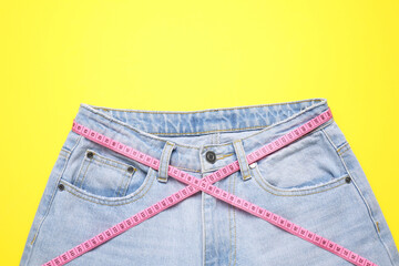 Jeans and measuring tape on yellow background, top view