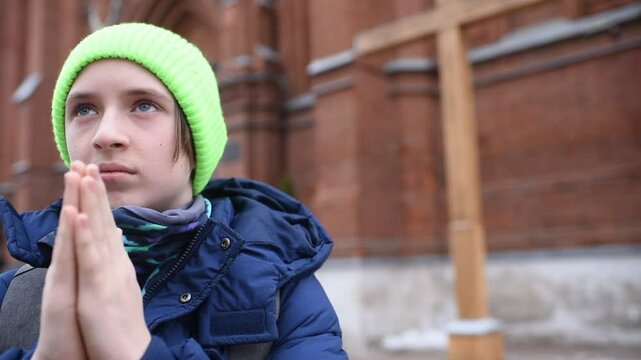12 year old boy in green hat and blue jacket prays in front of catholic church