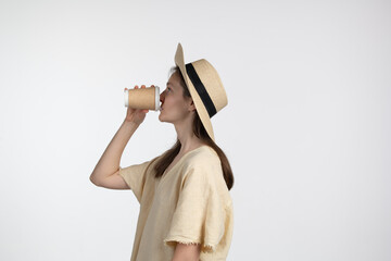 A woman stands in a minimalist environment, sipping coffee from a disposable cup. She wears a wide-brimmed straw hat and a simple, casual outfit, embodying relaxation and style
