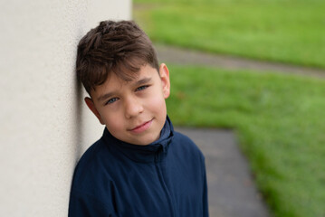 A young boy in a blue jacket leans against wall, looking thoughtful and relaxed. The outdoor background features a grassy area, conveying tranquil atmosphere. Boy resting after running