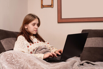 A young girl using a laptop on a cozy couch, wrapped in a warm blanket. She appears focused and...