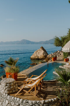A serene coastal retreat with a private pebble-lined pool overlooking the crystal-clear sea, surrounded by wooden decking, lounge chairs, and natural rock formations under the golden sun in T&uuml;rkiye