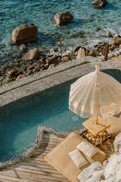 A serene coastal retreat with a private pebble-lined pool overlooking the crystal-clear sea, surrounded by wooden decking, lounge chairs, and natural rock formations under the golden sun in T&uuml;rkiye