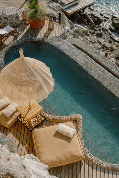 A serene coastal retreat with a private pebble-lined pool overlooking the crystal-clear sea, surrounded by wooden decking, lounge chairs, and natural rock formations under the golden sun in T&uuml;rkiye
