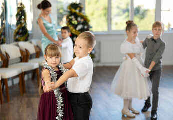 Enthusiastic preteen children, girls and boys in elegant outfits practicing partner dance moves in studio adorned with Christmas decorations, under guidance of female teacher