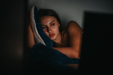 A young woman lies in bed at night, appearing emotional and deep in thought. The dim light enhances...