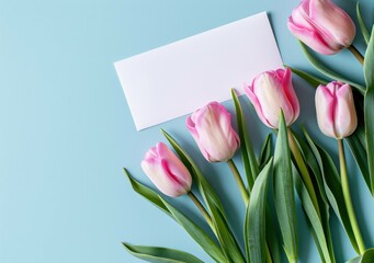 Fototapeta premium Beautiful pink tulips with green leaves arranged on a light blue background, accompanied by a blank card for personalized messages. Floral and greeting concept.