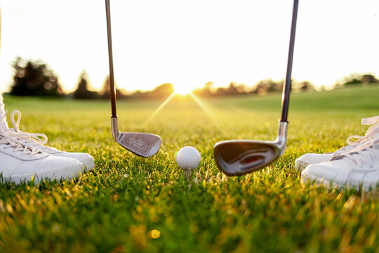 young sports couple in uniform playing golf on the course with clubs and ball on green grass, close-up of golfers' legs in white shoes at sunset - Powered by Adobe