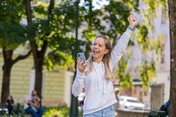Fototapeta premium Happy Caucasian young woman using smartphone celebrating win good message news, lottery casino app jackpot victory, online outdoors. Attractive female tourist teenager clenching fists on city street.