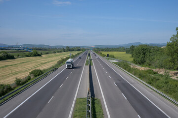 A highway stretching into the horizon, surrounded by green fields and trees under a clear blue sky.