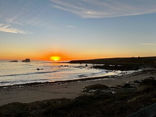 A serene sunset over a coastal beach with seals resting on the shore, rocky outcrops in the water, and a clear sky with wispy clouds.